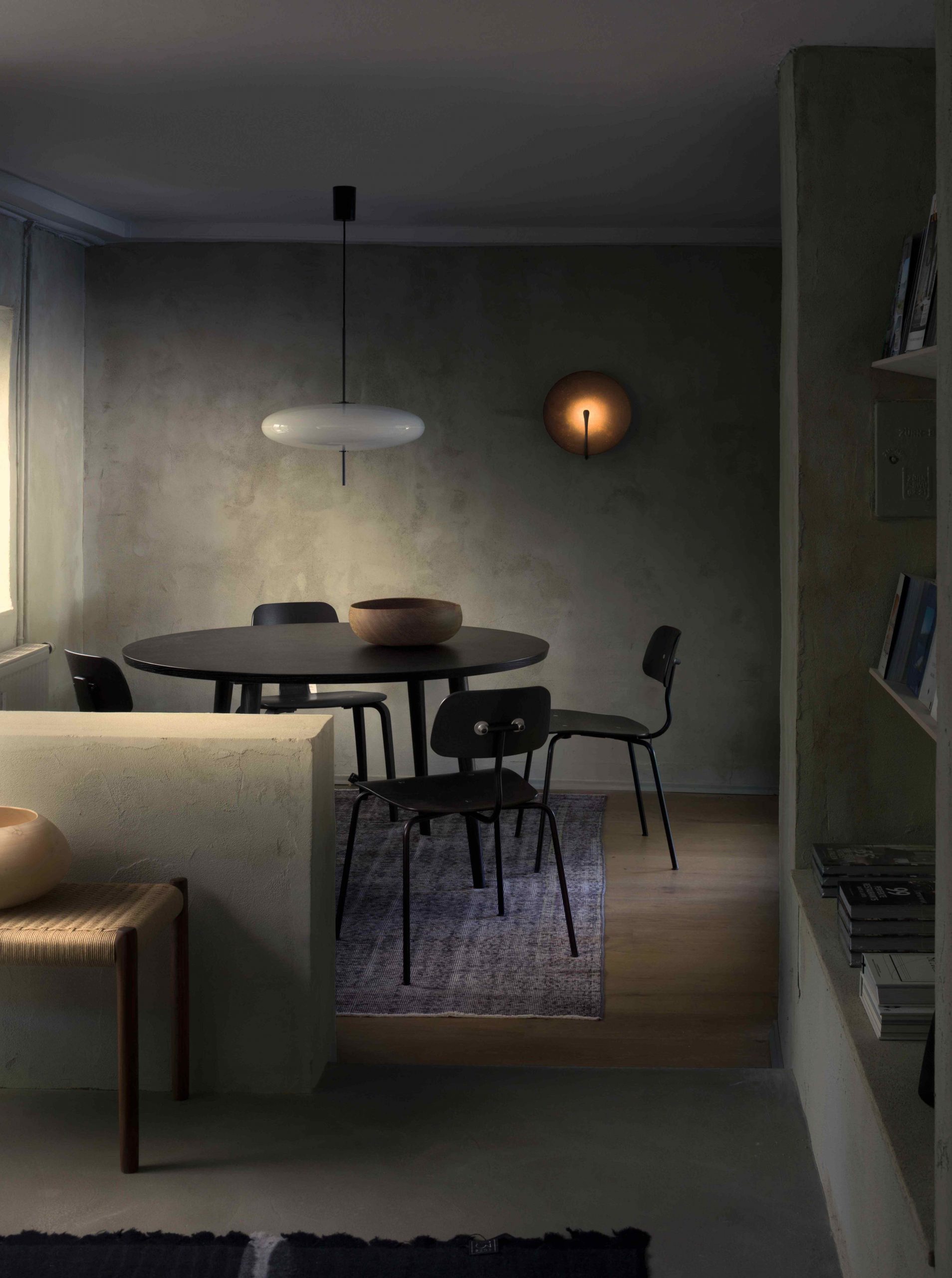 Stimmungsvoll beleuchteter PEAM Showroom mit rundem schwarzen Esstisch, klassischen Holzschalen, minimalistischer Pendelleuchte und Wandlampe vor gespachtelter Kalkputzwand – ruhiges Interior Design mit skulpturaler Wirkung