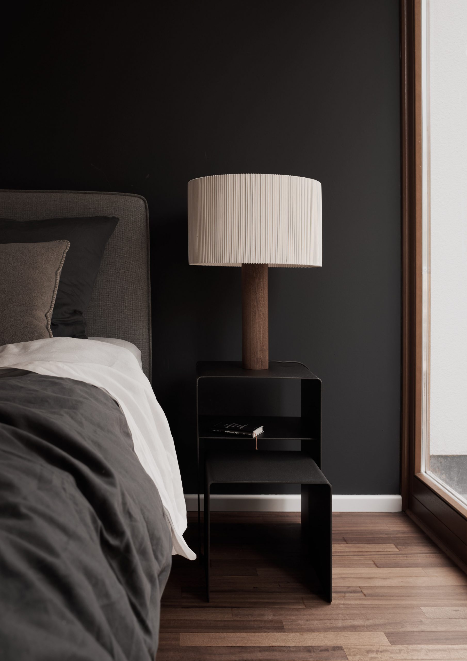 Minimalistisches Schlafzimmer mit dunkler Wand, modernem Nachttisch aus Metall, Tischleuchte Moragas mit Holzfuß und gefaltetem Lampenschirm, Bett mit grauer Bettwäsche auf Echtholzboden