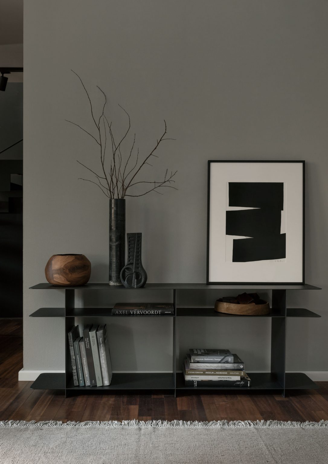 Modernes, offenes Regal in schwarzem Metall mit Designobjekten, Kunstbüchern und minimalistischer Dekoration vor einer grau gestrichenen Wand auf dunklem Holzfußboden