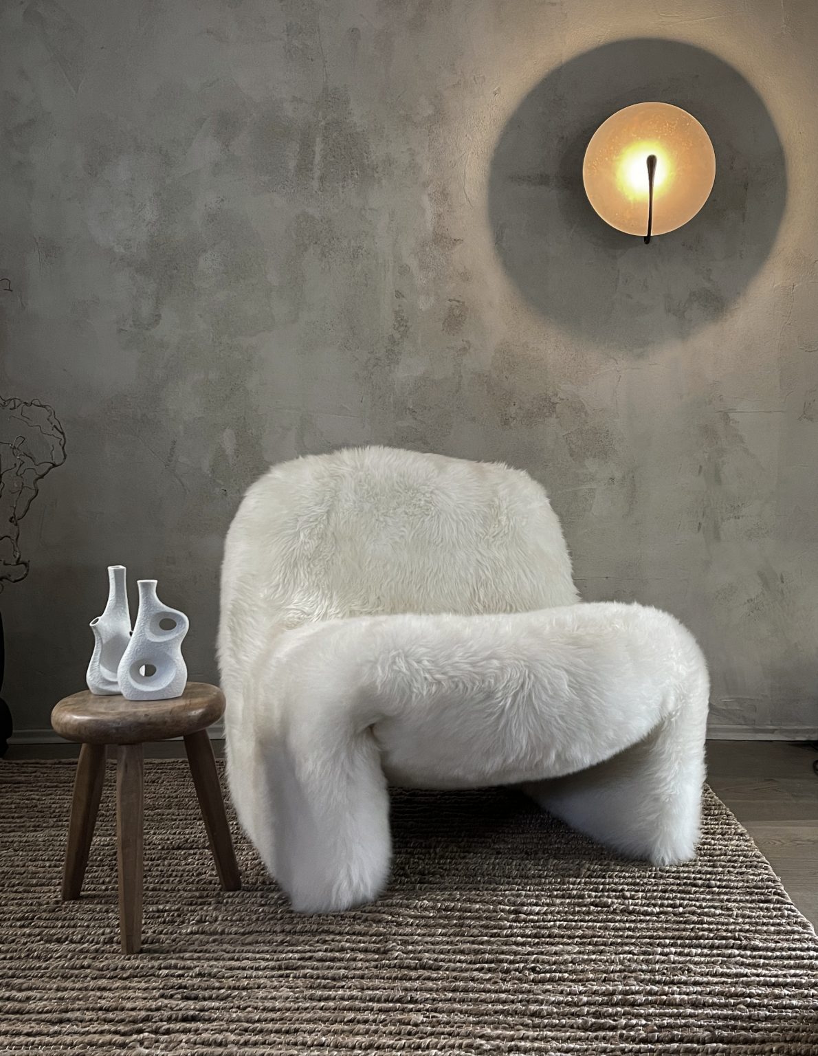 Skulpturalen Weißer designer sessel mit Schaffell bezogen und runder Designer Wandleuchte Vintage Holzbockes mit zwei Vintagevasen vor einer Kalkputzwänden im PEAM Showroom auf einem naturfarbenem Hanfteppich